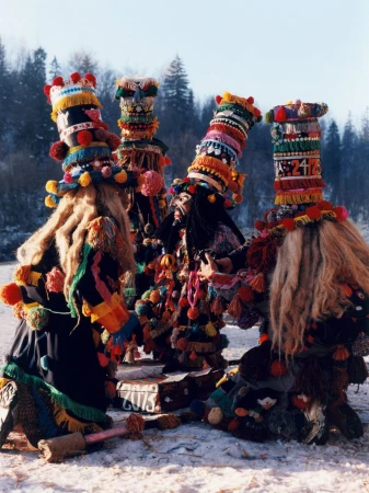 Five people in traditional carnival costumes with tall hats, tassels, and long fur, standing in a snowy field with trees in the background.