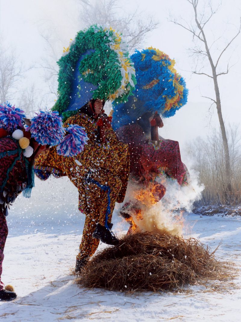 Performers in colourful costumes and elaborate headdresses participate in a winter ritual, standing by a burning pile of sticks.