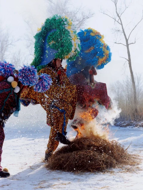 Performers in colourful costumes and elaborate headdresses participate in a winter ritual, standing by a burning pile of sticks.