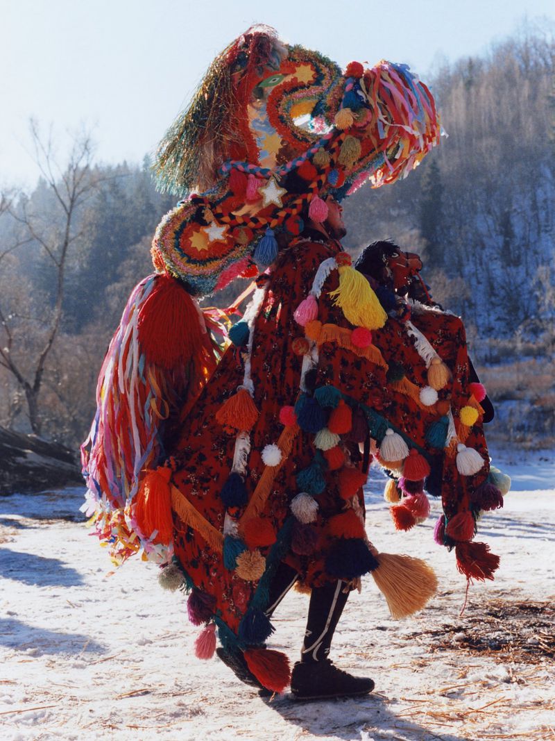 Person in colourful, elaborate costume with pom-poms and tassels stands in a snowy outdoor setting against a backdrop of trees and hill.