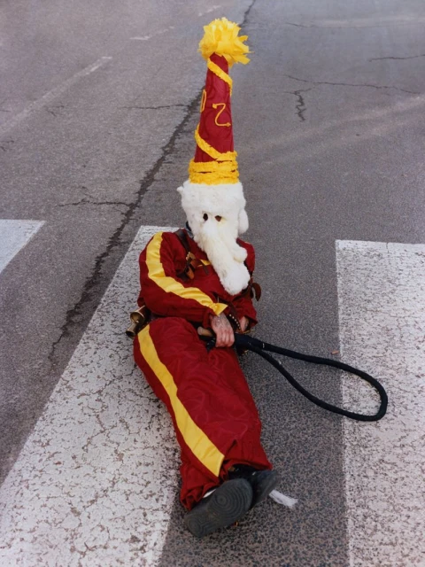 A person in a festive red and yellow jester costume sits on a crosswalk, wearing a tall pointed hat and a white mask.