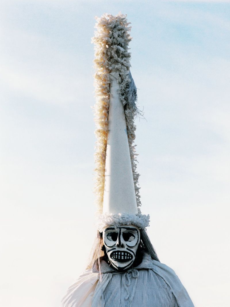 Person wearing a tall, conical, white cap and painted mask, white robe against a light blue sky.