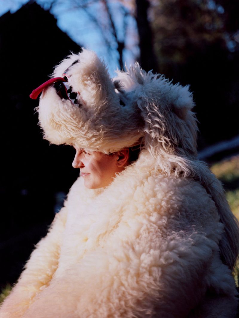 Adult female in a fur-like costume with a hat, looking to the left in an outdoor, sunlit environment with trees in the background.