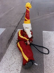 A person sitting on a paved pedestrian crossing wearing a red and yellow jumpsuit, white mask, and tall cone hat.
