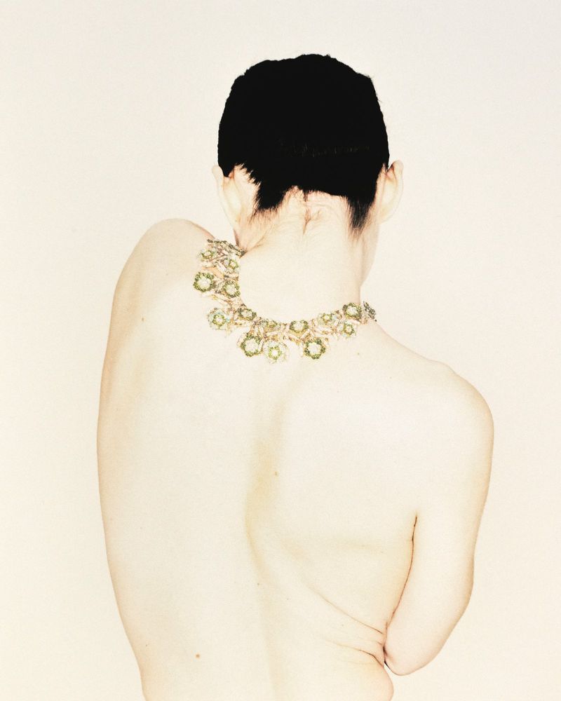 Back view of a bare-backed person with a short, dark haircut and a detailed gold and green necklace against a pale, neutral background.