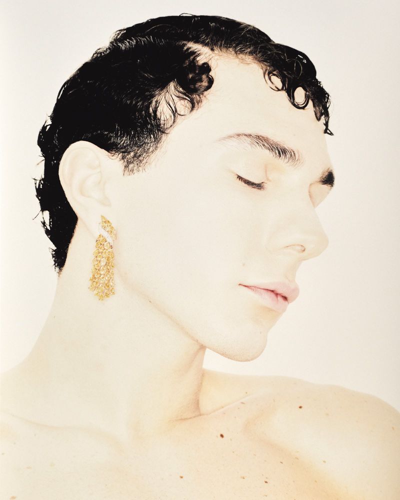 Elegant portrait of a young adult with black hair, eyes closed, and golden tassel earrings, set against a soft, light background.