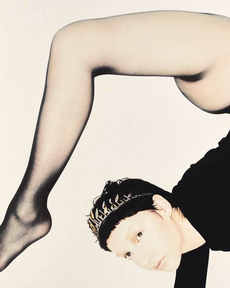 Woman in black top and tights poses with legs in the air against a white backdrop in a fashion photography studio shot.