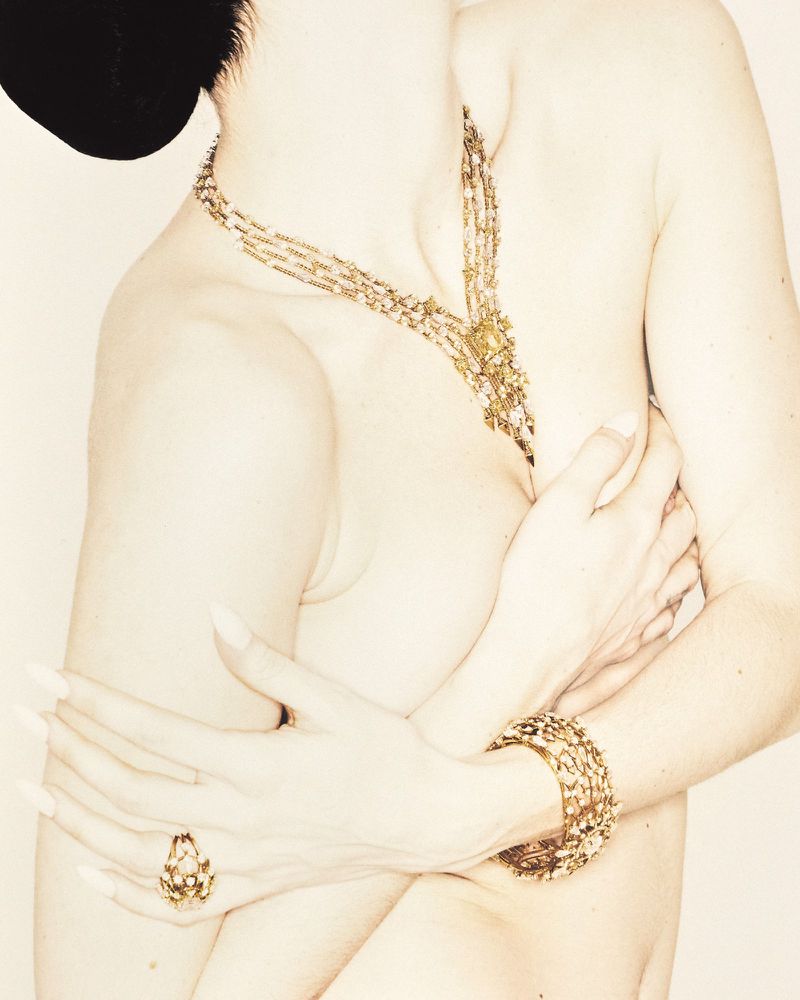 Close-up portrait of a model wearing a gold layered necklace, bracelet, and ring against a neutral background.