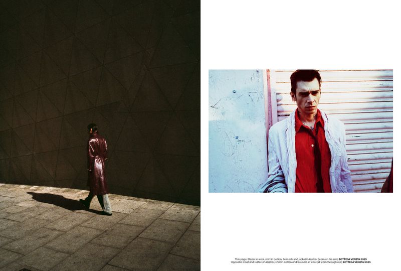 Editorial fashion shot of a man in a leather coat walking past a textured wall and another man in a red shirt leaning against a wall.