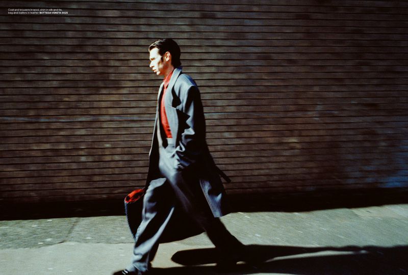 Fashion editorial shot of a man in a suit coat walking in an urban environment with a brick wall, showcasing fashion and style.