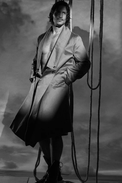 Monochrome portrait of a handsome adult man in a jacket, partially unbuttoned, against a backdrop of ropes and a cloudy sky.