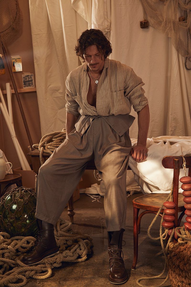 Masculine fashion shot: A man poses indoors, seated on a chair in a striped shirt, grey trousers, and brown boots, amid fishing nets and ropes.