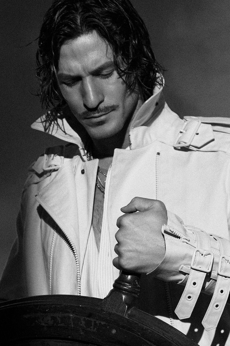Monochrome portrait of a stylish man with long hair, a mustache, and a buckled white leather jacket holding a ship's helm.