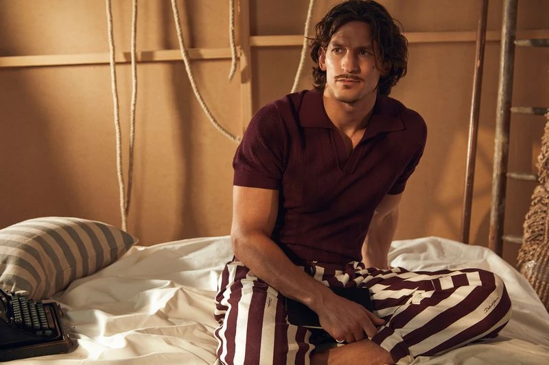 Adult male in a burgundy collared shirt and striped pants sits on a bed indoors with a typewriter in a lifestyle fashion shot.