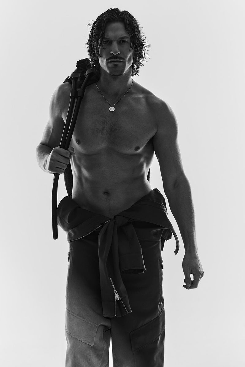 Monochrome portrait of a shirtless adult male with a backpack, showcasing fitness and fashion against a bright studio backdrop.