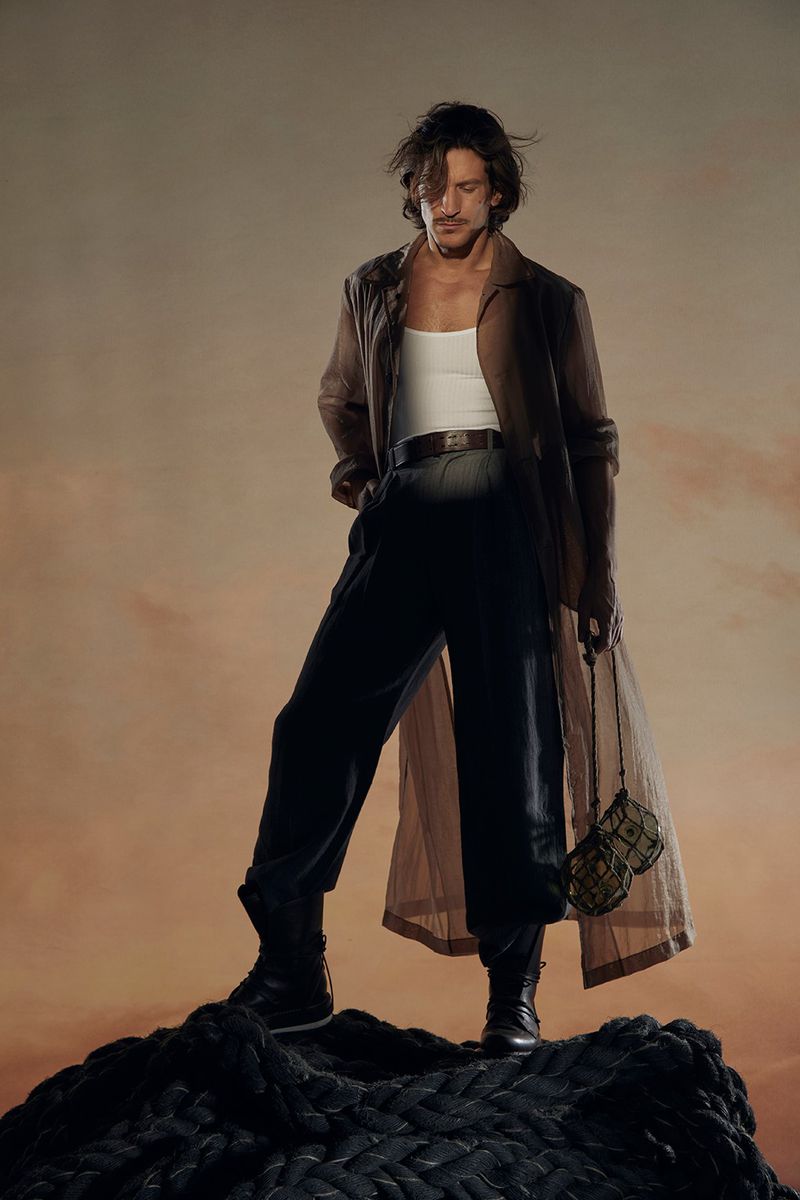 A stylish man in a brown coat poses on a black rope structure against an orange gradient background, captured in a studio setting.