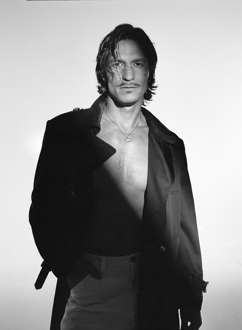 Portrait of a man wearing a dark coat and pendant necklace against a white background