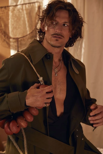 Portrait of a man with long hair and a mustache wearing an unbuttoned green trench coat, necklace, and rope across his shoulder.