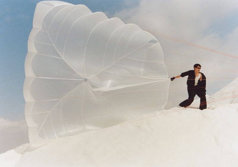 A man in sunglasses and dark clothing holds a large white parachute-like structure on a snow-covered landscape against a bright sky backdrop.