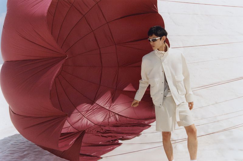 A stylish Asian man in a white suit stands in the desert by a large burgundy parachute, wearing sunglasses and looking away from the camera.