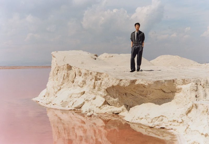 A well-dressed man stands atop a sand dune overlooking a pink salt lake on a cloudy day, creating a surreal, fashion-forward image.