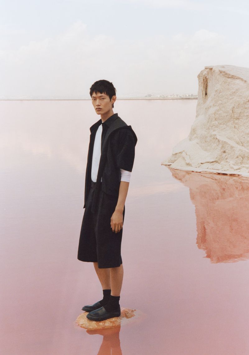 A person stands in black jacket, shorts, white shirt on a small patch in a pink lake, posing in a tranquil outdoor scene near rocks.