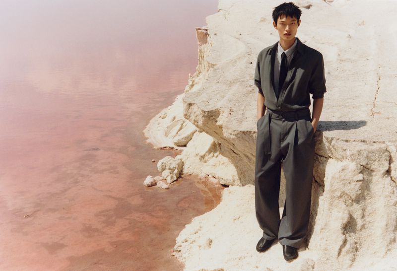 Adult man in gray suit poses on rocky cliff overlooking pink lake in outdoor fashion shot with natural light, medium shot, editorial style.