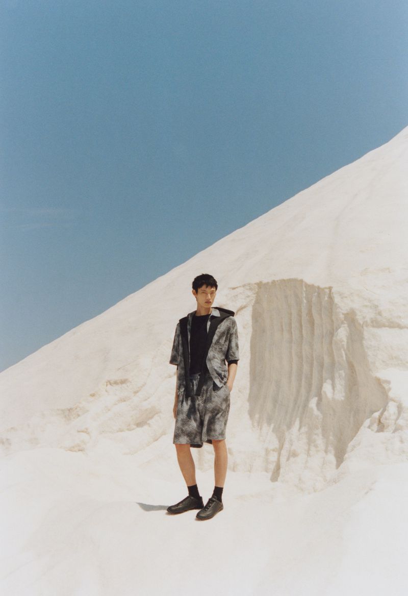 Asian man in a tie-dye outfit stands in a bright landscape with a pile of white rocks under a clear blue sky outdoors.
