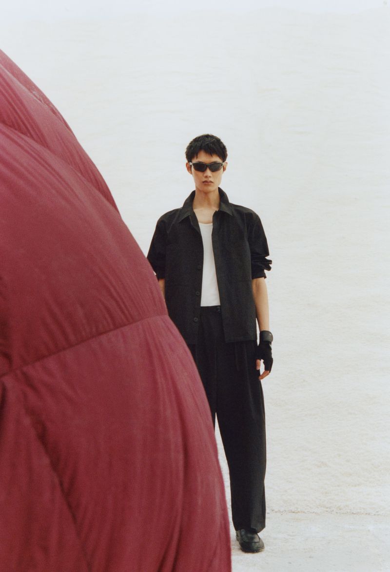 Asian male model in sunglasses and dark jacket posing next to large red inflated object against a light background in an outdoor setting.