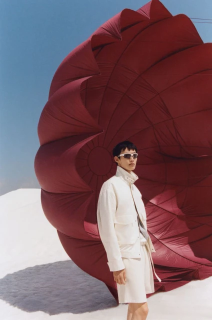 Fashion shot of person wearing white jacket and shorts standing near a large, red parachute on sand against a clear sky.