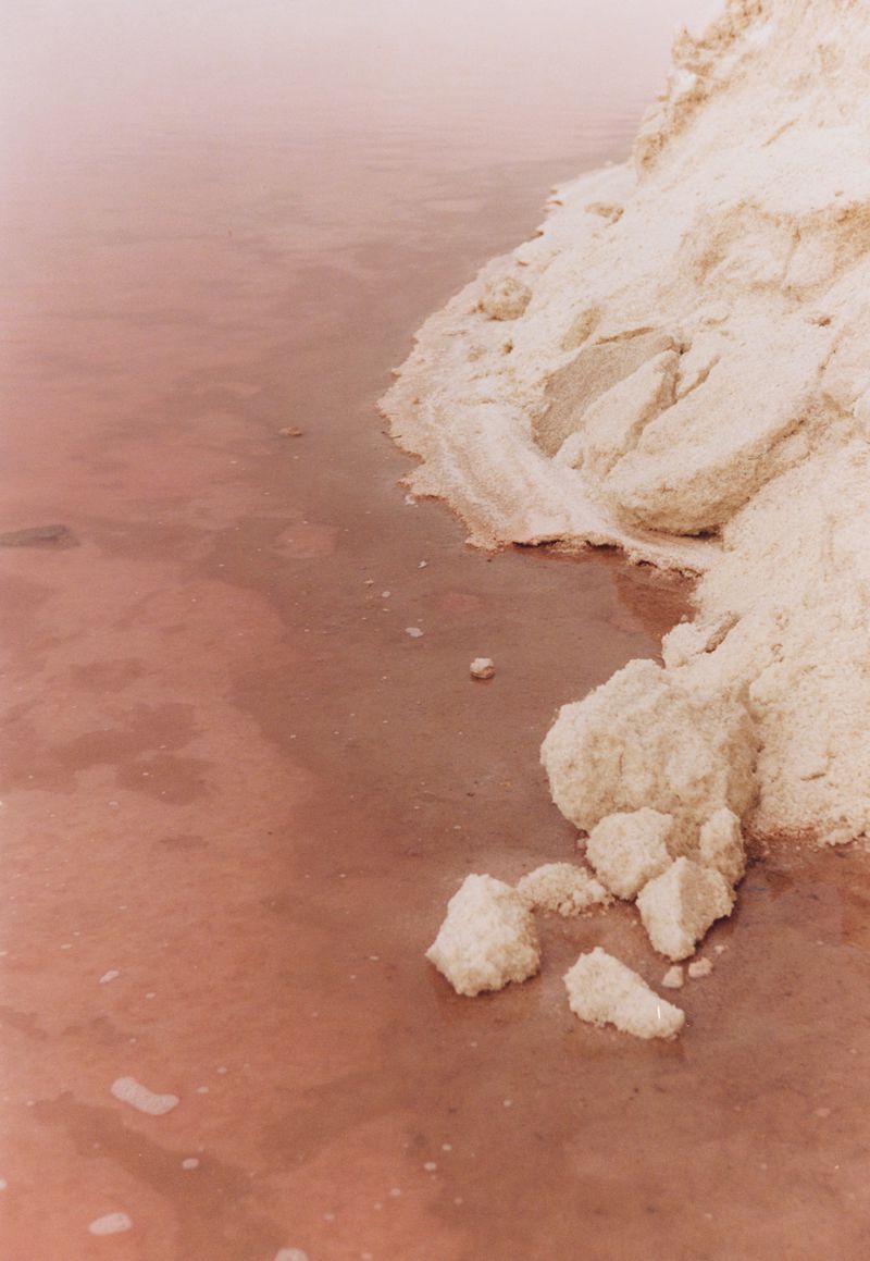 Pink salt lake with white mineral deposits along the shore, creating a surreal and minimalist nature landscape with muted colors.