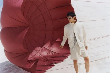 Model wearing cream technical jacket and shorts holding strings of a large red parachute on snowy ground
