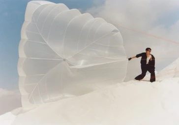 Model pulling a large white parachute on a white sandy dune under an open sky for a Zara technical wardrobe fashion campaign.