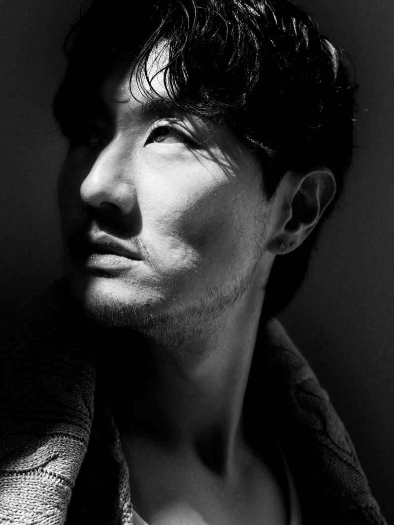 Black and white portrait of an adult male with dark hair and a light beard, looking up to the light, wearing a knit sweater.