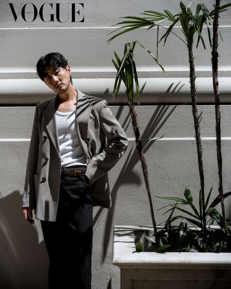 Editorial fashion shot of a man in a grey suit jacket and black jeans standing near a decorative planter under bright natural light.