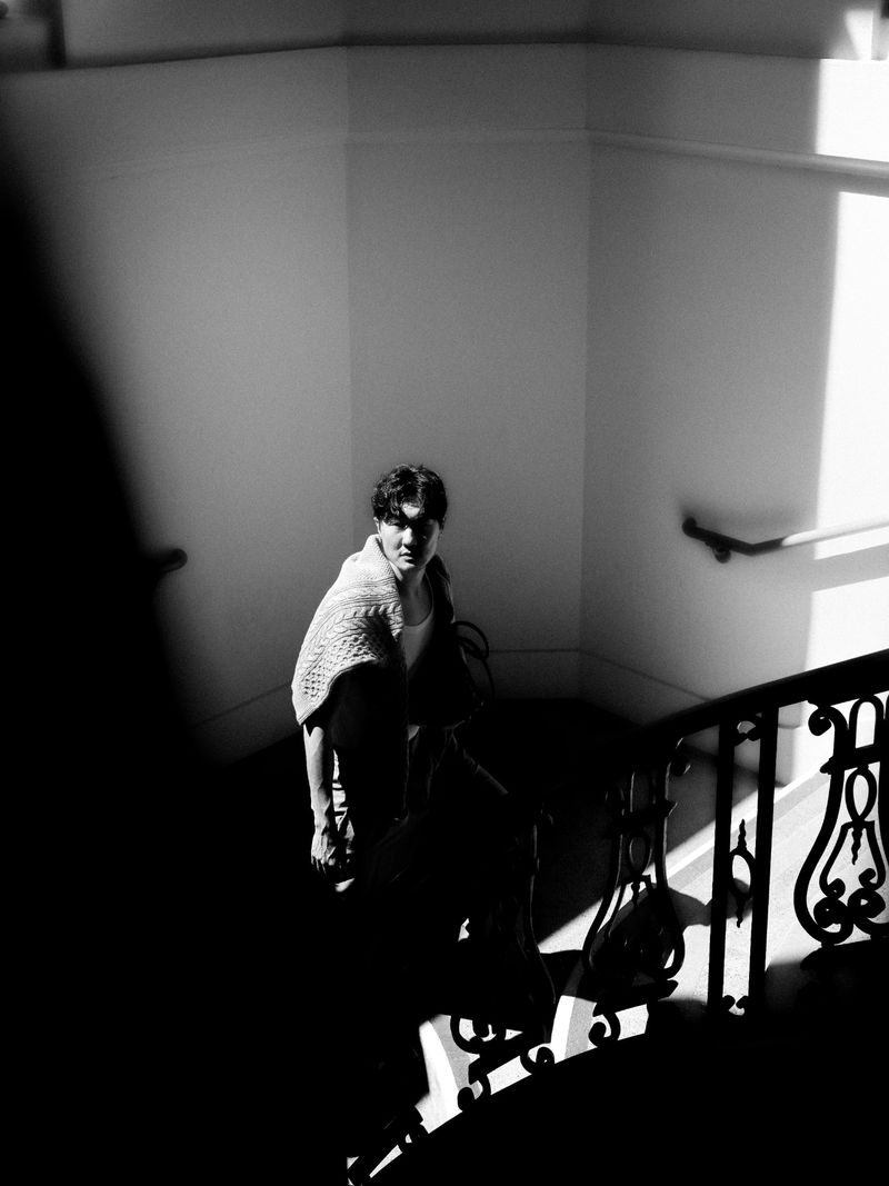 Black and white portrait of a woman standing on a staircase with ornate metal railings and bright light coming from a window.