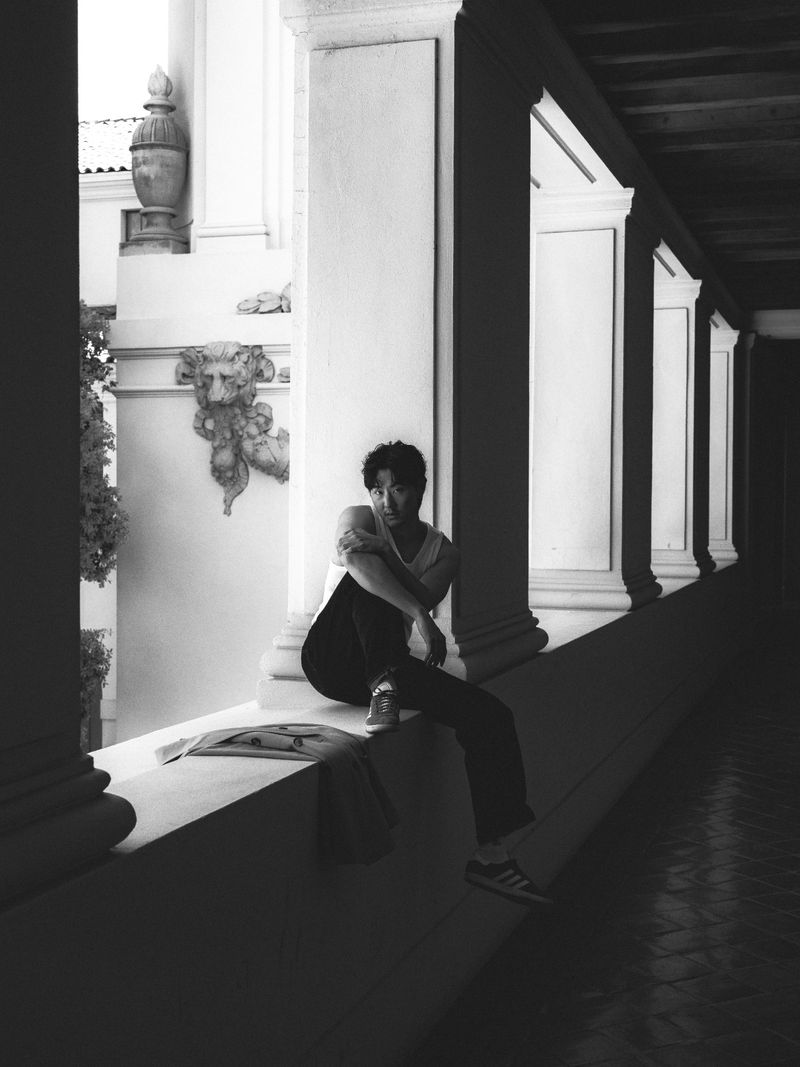 Young man in casual clothes sits on a ledge with a document in a colonnade. The setting has classic architecture in monochrome.