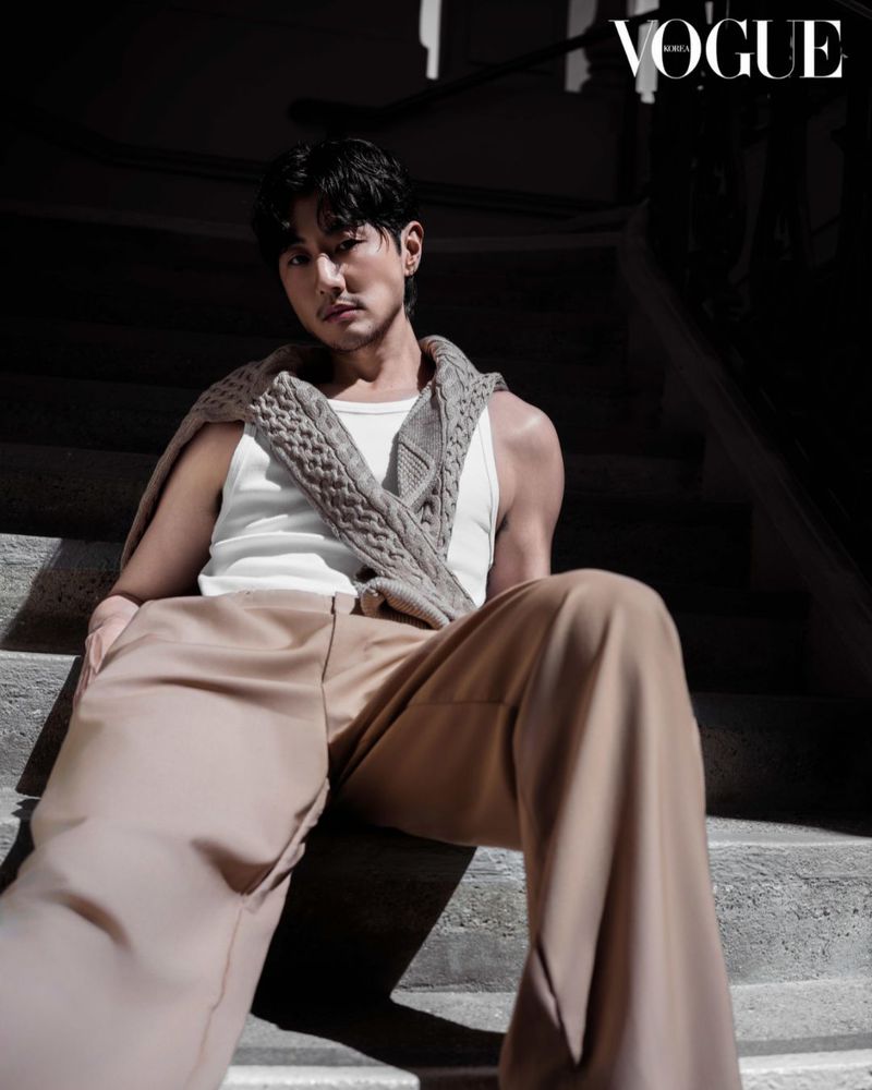 Stylish adult male poses sitting on stairs wearing a white tank top and tan pants, with a gray sweater draped over his shoulders.