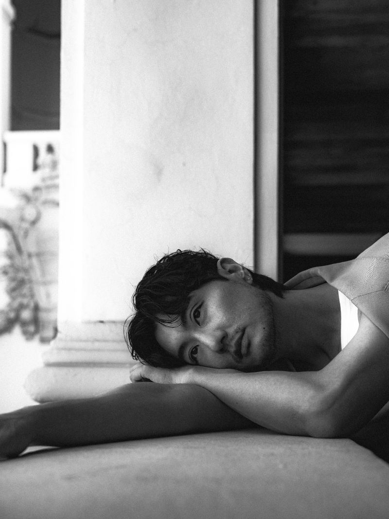 Portrait of David Yi resting his head on his arms outdoors against a classical architectural column, shot in black and white.