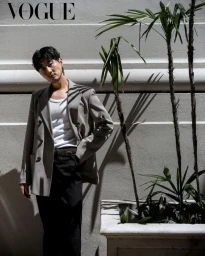 Man wearing a grey blazer, white tank top, and dark trousers standing against a textured wall with potted palm trees