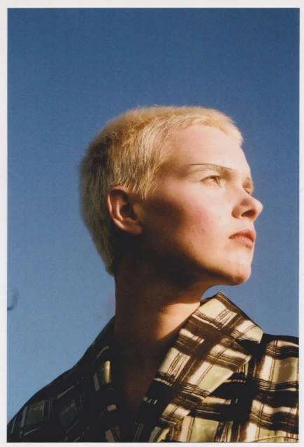 Portrait of a person with short blonde hair, wearing a patterned blazer, looking upwards against a clear blue sky.