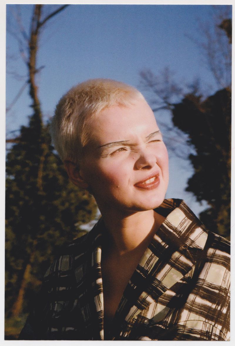 Woman with a buzzcut poses in a brown and beige plaid jacket with a forest and blue sky in the soft-focus background.