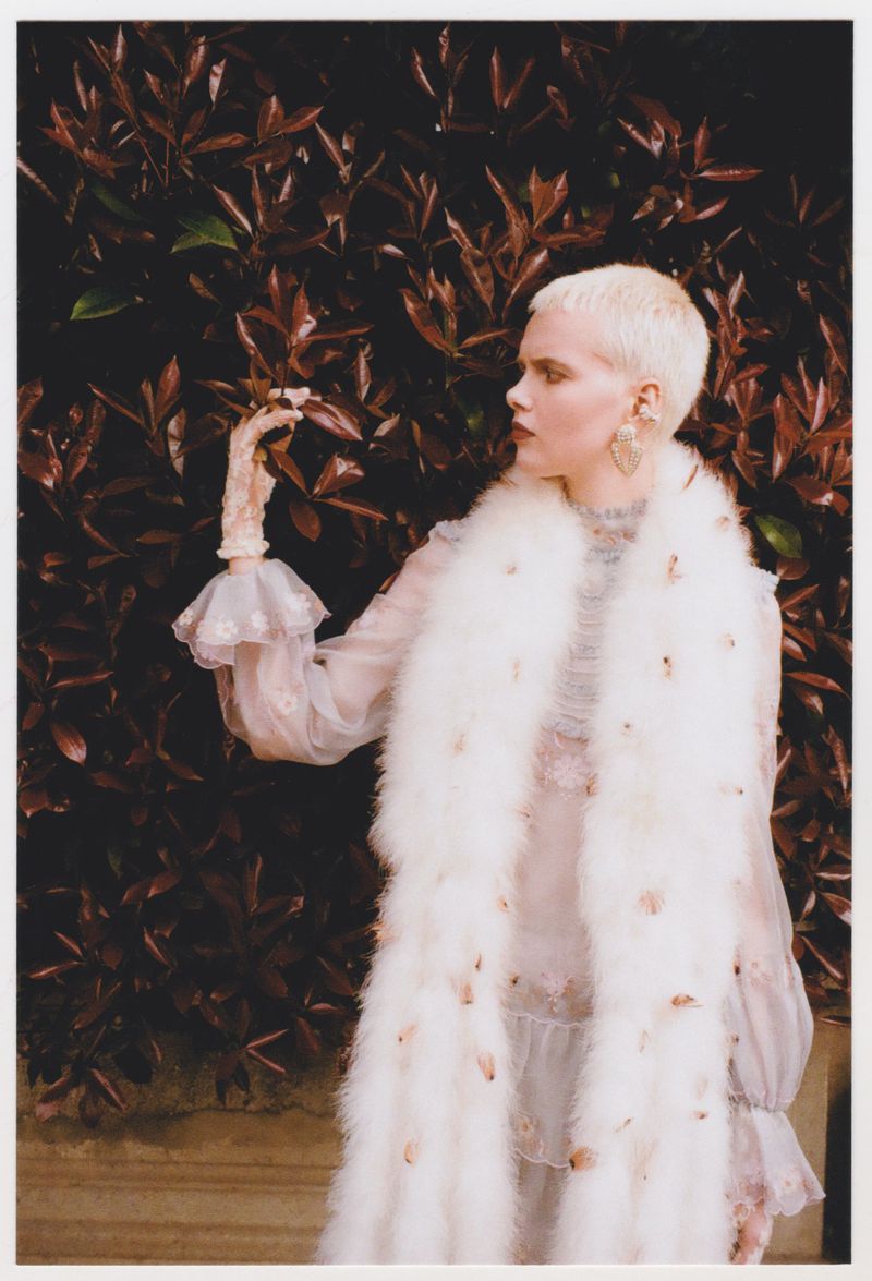 Woman in a white fur stole and sheer dress poses near reddish-brown foliage, looking off to the side with delicate hand gestures.