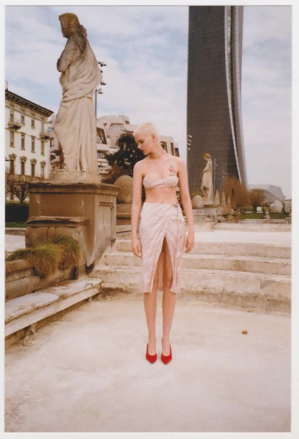 Fashion shot of a model in a pale outfit and red high heels standing in front of statuary and a modern skyscraper in a European city.