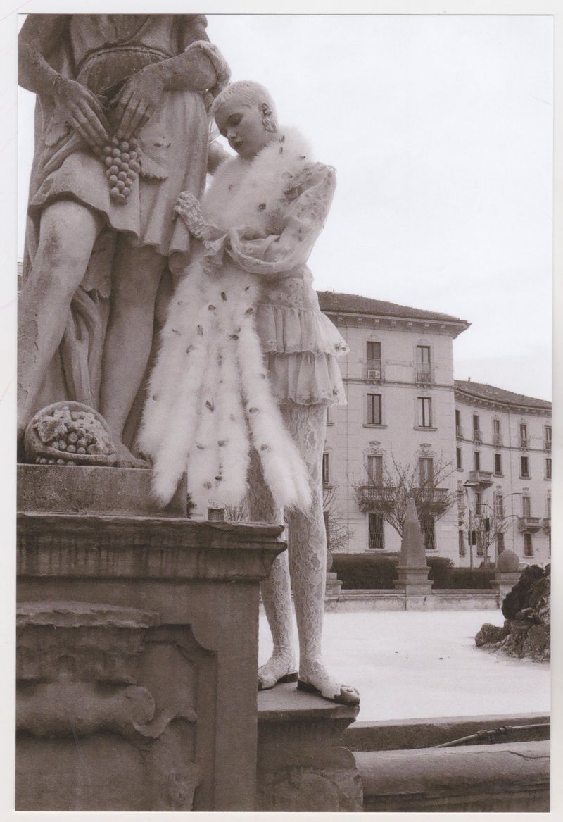 Two statues, one adorned with feathers, stand outdoors. A building and bare trees create a backdrop. The scene is monochrome.