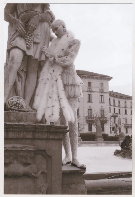Two statues, one adorned with feathers, stand outdoors. A building and bare trees create a backdrop. The scene is monochrome.