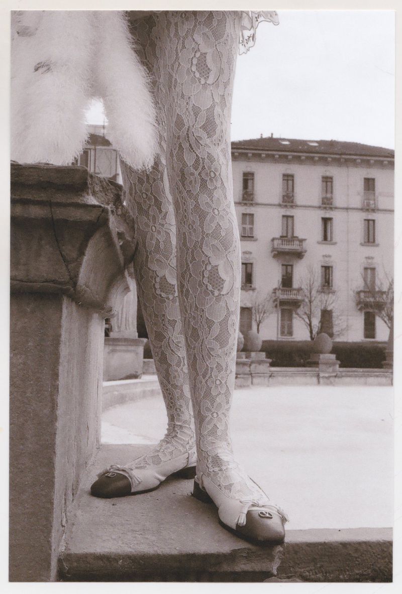 Close-up of a woman's legs in floral lace leggings, paired with flats, outside near a building in grayscale.