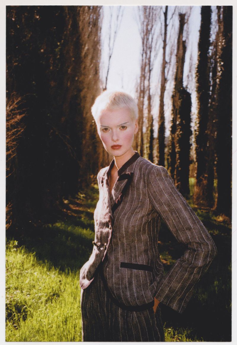 An adult female model with short white hair poses outdoors in a pinstripe suit, standing in a grassy area with trees in the background.