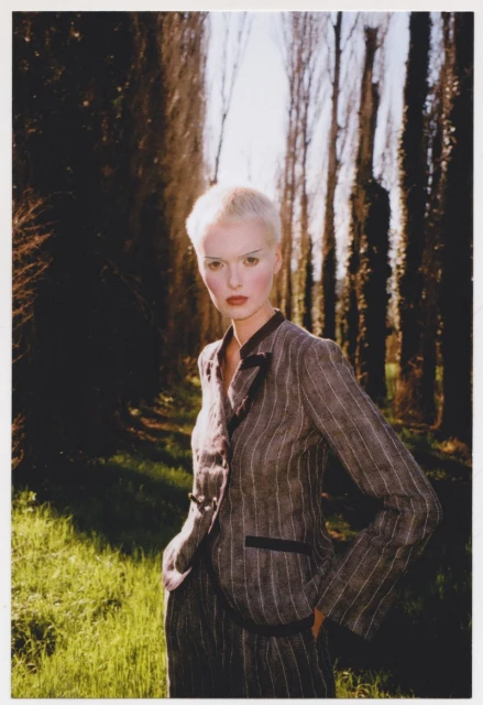 An adult female model with short white hair poses outdoors in a pinstripe suit, standing in a grassy area with trees in the background.