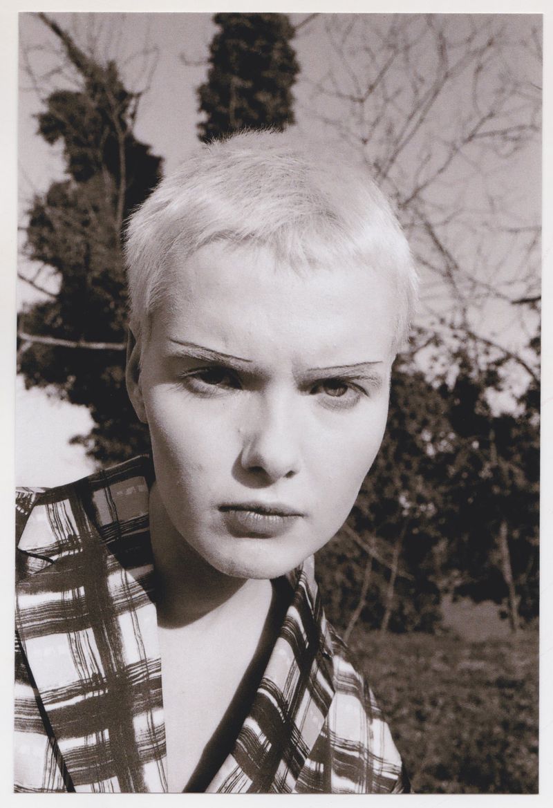 Portrait of an androgynous adult with short platinum hair, wearing a plaid jacket and striking a pose against an outdoor background of trees.